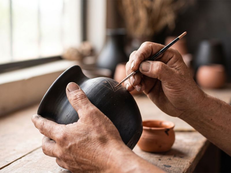 Artisan hands hand-painting fine detail onto a black clay pottery vessel