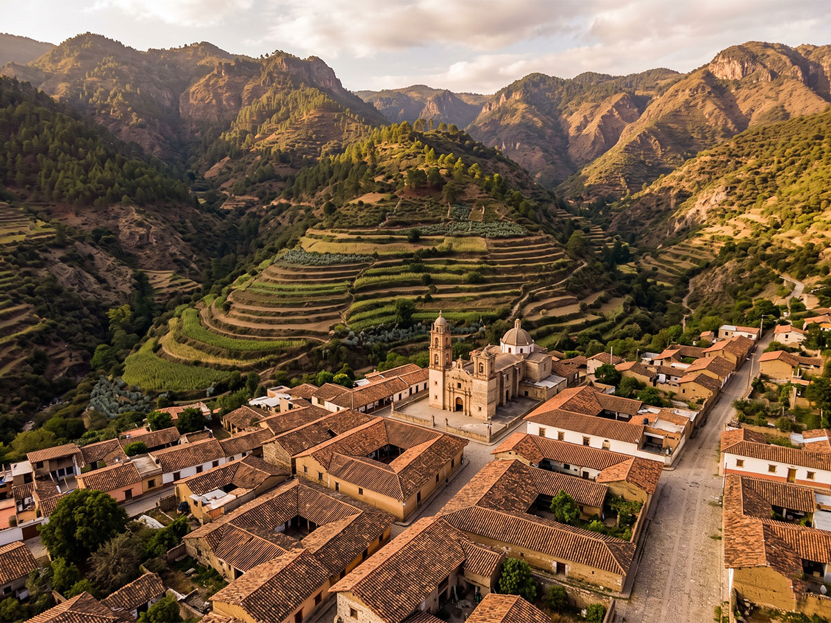 Aerial view of a traditional Mexican village in a valley surrounded by mountains, terracotta rooftops visible among trees