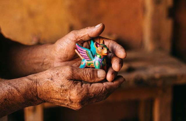 A pair of weathered hands cradling a small hand-painted ceramic figure, warm studio light